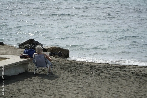 Fototapeta Ostia, Rome, Italy - October 19, 2025, a lady from behind sunbathes on the beach on a folding chair on a beautiful October day, called ottobrate romane in Rome.