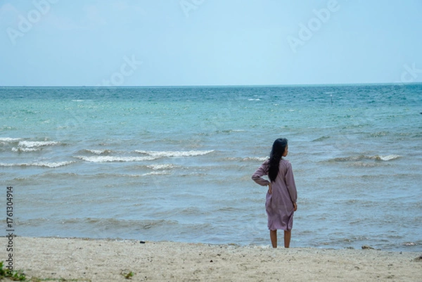 Fototapeta long haired asian woman wearing light purple clothes with long sleeves standing facing the sea on the beach with bright sky background for advertis