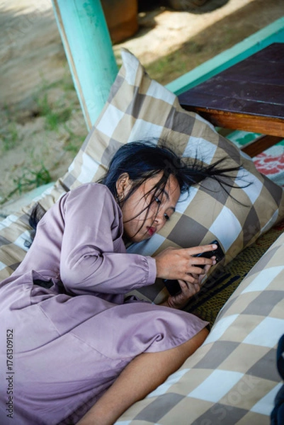 Fototapeta A view of an Asian woman with long black hair lying on a checkered patterned pillow while holding a cellphone with both hands and with a serious expre