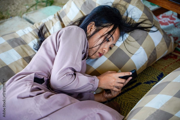 Fototapeta A view of an Asian woman with long black hair lying on a checkered patterned pillow while holding a cellphone with both hands and with a serious expre