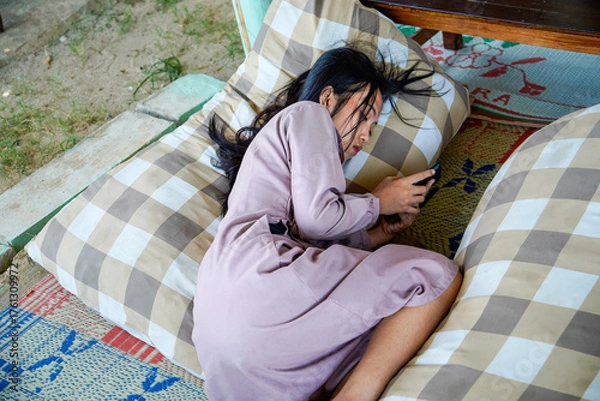 Fototapeta A view of an Asian woman with long black hair lying on a checkered patterned pillow while holding a cellphone with both hands and with a serious expre