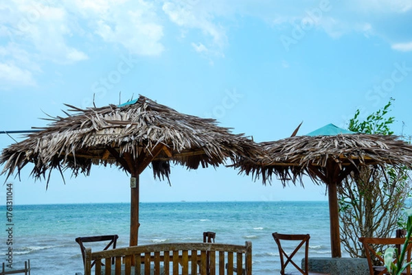 Fototapeta view of the seating area with large straw umbrellas with a backdrop of a beachfront view with bright blue skies and a horizon that reveals land in the distance.