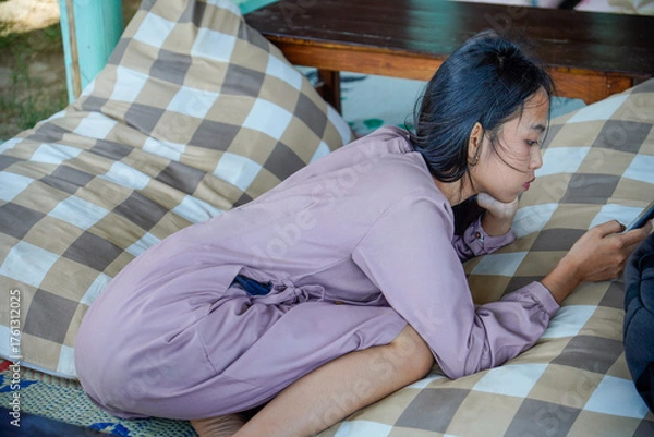 Fototapeta A view of an Asian woman with long black hair lying on a checkered patterned pillow while holding a cellphone with both hands and with a serious expre