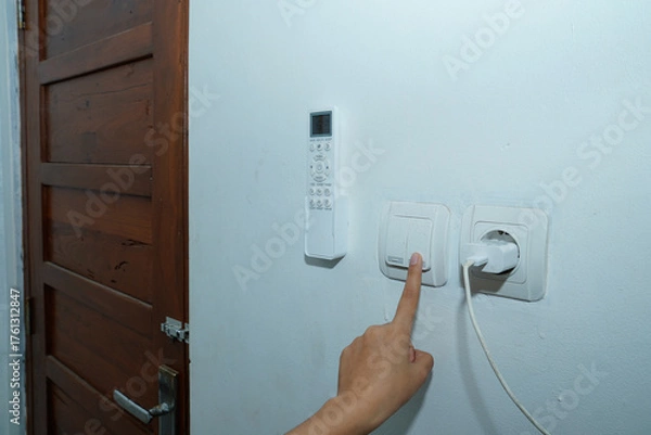 Fototapeta hand trying to turn on the light switch on the white wall with the AC remote control on the left and the socket with the charger cable connected to the right.
