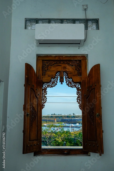 Fototapeta Wooden windows with carved motifs that open to reveal the outside view of water, trees and bright blue sky.
