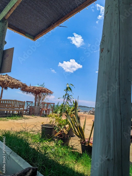 Fototapeta a view of a bright blue sky on a sandy beach with a few wooden chairs and straw umbrellas from behind the window frame.