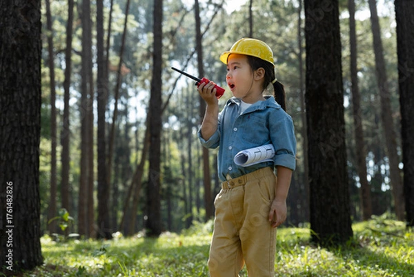 Fototapeta Little girl in safety helmet holding blueprints and talking on a walkie-talkie.