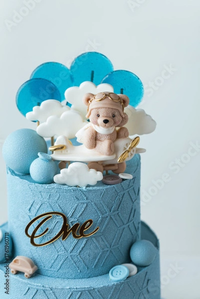 Fototapeta Big two tier birthday cake with blue chocolate frosting decorated with teddy bear flying among the clouds on the plane figurine. Beautiful birthday cake for a little one year old boy. White background