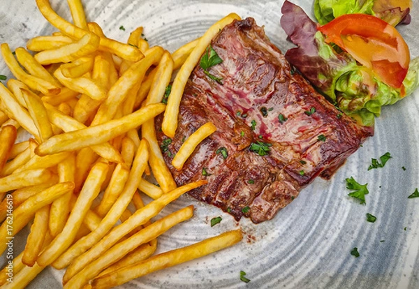 Obraz A large steak frites plate, featuring a well-seared skirt steak piled next to a generous portion of golden french fries. The meal is garnished with parsley and a simple salad of lettuce and tomato