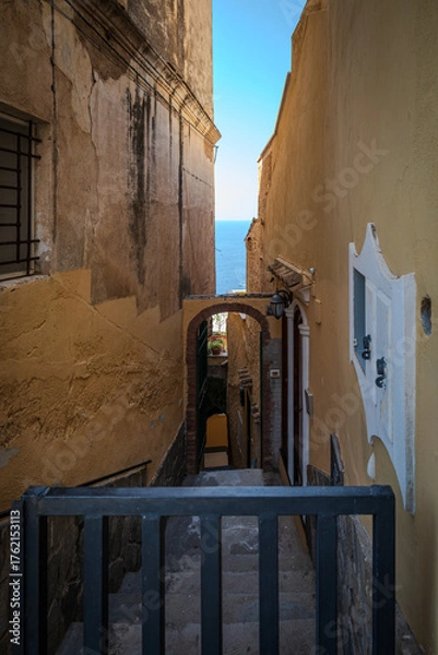 Fototapeta Narrow downhill lane with stairs between cliffside houses in Positano, Italy, with a sliver of blue sky and sea visible in the distance