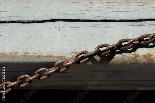 Fototapeta Rusted metal chain lying on weathered wooden surface. Old industrial chain casting shadow on faded paint.