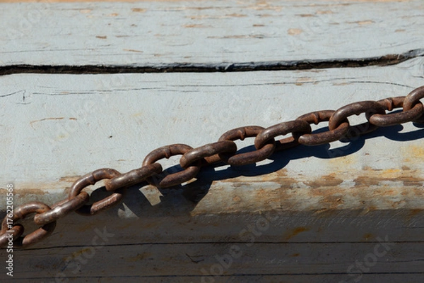 Fototapeta Rusted metal chain lying on weathered wooden surface. Old industrial chain casting shadow on faded paint.