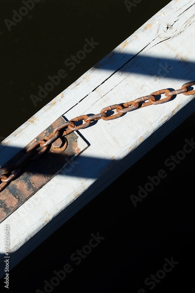 Fototapeta Rusty chain attached to weathered wooden beam over dark water. Old metal chain and shadow detail on sunlit dock surface.
