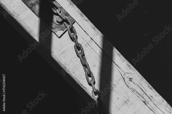 Fototapeta Rusty chain attached to weathered wooden beam over dark water. Old metal chain and shadow detail on sunlit dock surface.