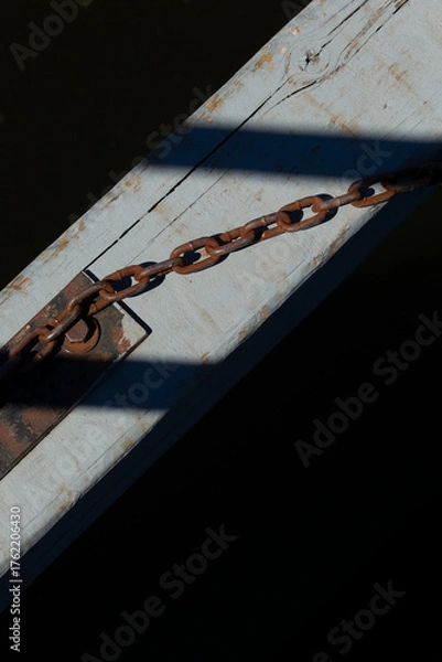 Fototapeta Rusty chain attached to weathered wooden beam over dark water. Old metal chain and shadow detail on sunlit dock surface.