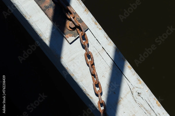 Fototapeta Rusty chain attached to weathered wooden beam over dark water. Old metal chain and shadow detail on sunlit dock surface.