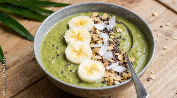 Fototapeta A healthy green smoothie bowl topped with banana slices, granola, and coconut flakes. The bowl is placed on a wooden surface with green leaves in the background.