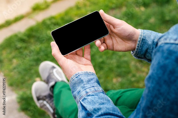 Fototapeta Man use his Mobile Phone outdoor, close up. Mobile phone in hands a young hipster business man in denim shirt and green jeans on the background of green grass.