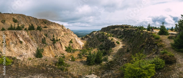 Fototapeta Warstwowy krajobraz kamieniołomu ze skalistymi klifami i rozproszonymi zielonymi drzewami pod zachmurzonym niebem 
