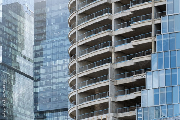 Fototapeta curved skyscraper under construction.  contrasts raw stages of vertical development next to a completed modern facade. office building with a grid of blue glass windows against the exposed concrete