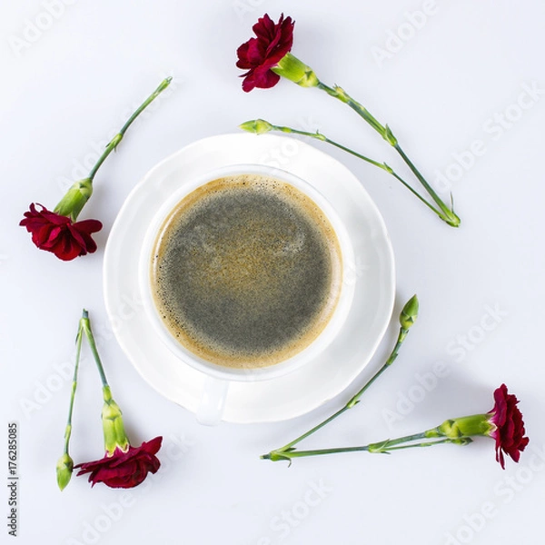 Fototapeta A cup of coffe in the centre and red carantions around on white