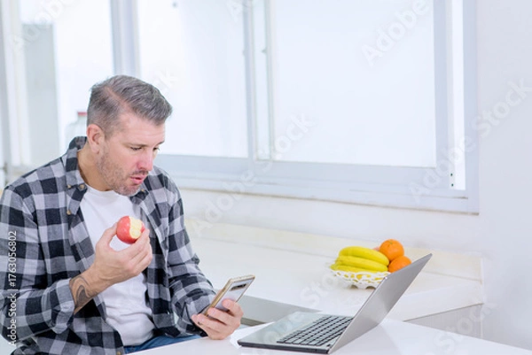 Fototapeta Multitasking Man Eating Healthy Apple and Checking Smartphone While Working on Laptop in Modern Kitchen