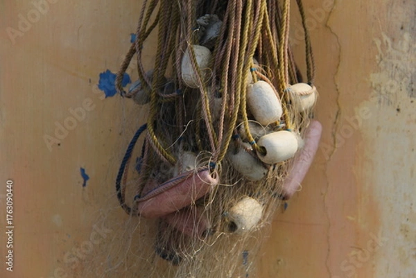 Fototapeta Close-up of fishing net with white and pink floats hanging against a weathered wall. Ideal for maritime, fishing, rustic, or traditional-themed visual conten