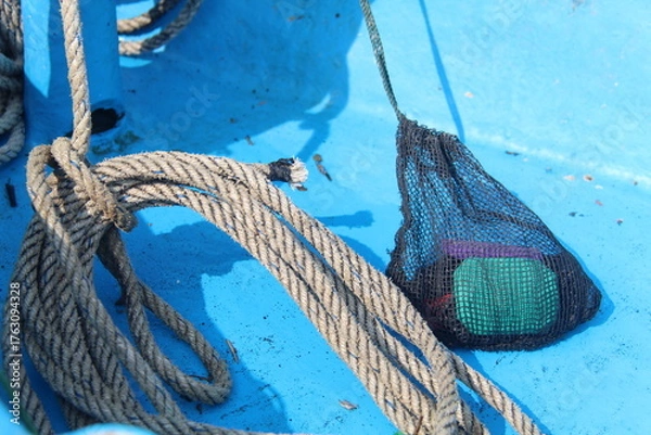 Fototapeta Close-up of a thick marine rope and a small fishing net on a blue boat deck under sunlight. Detail of nautical equipment used by fishermen on a traditional wooden boat.