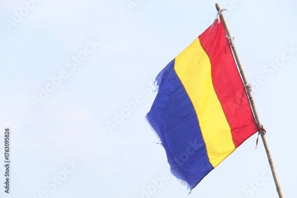 Fototapeta Romanian flag with vertical blue, yellow, and red stripes waving on a pole against a clear sky. Ideal for national, cultural, travel, or identity-themed visual content