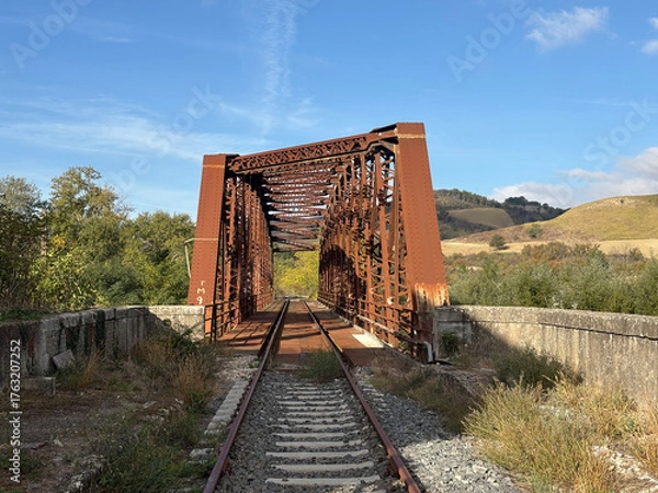 Obraz An old, rusty iron railway bridge spans a valley surrounded by nature, amidst golden hills and a clear sky.