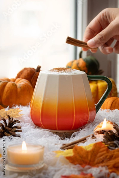 Fototapeta A cup of coffee on a pile of books against window view with cozy atmosphere and autumn ambiance, with Jack Be Little squash pumpkin and autumn leaves, while hand hold cinnamon putting over a cup.
