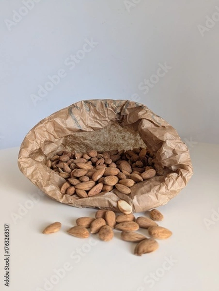 Fototapeta Raw almonds pouring from a brown craft paper bag onto a bright white surface. Capturing natural, healthy movement.
