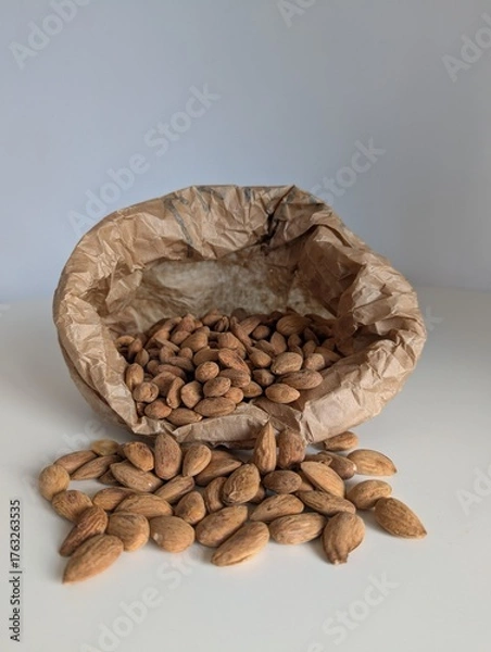 Fototapeta Organic almonds spilling from a rustic paper bag onto a clean white surface. A natural, wholesome food concept.