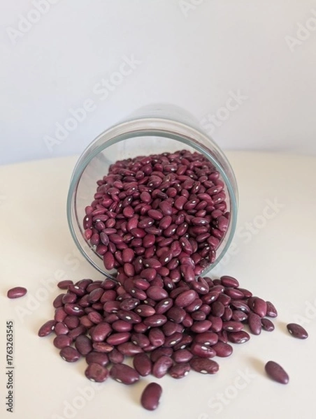Fototapeta A minimalist flat-lay of red beans poured from a clear glass jar onto a pure white surface. Simple and clean.