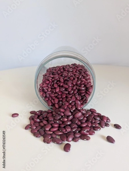 Fototapeta Red kidney beans pouring from a tilted glass jar onto a white surface. Simple, natural food ingredient.