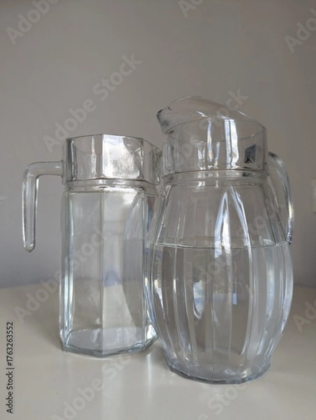 Fototapeta Two different shaped glass water jugs side by side on a white table. A simple, effective stock image.