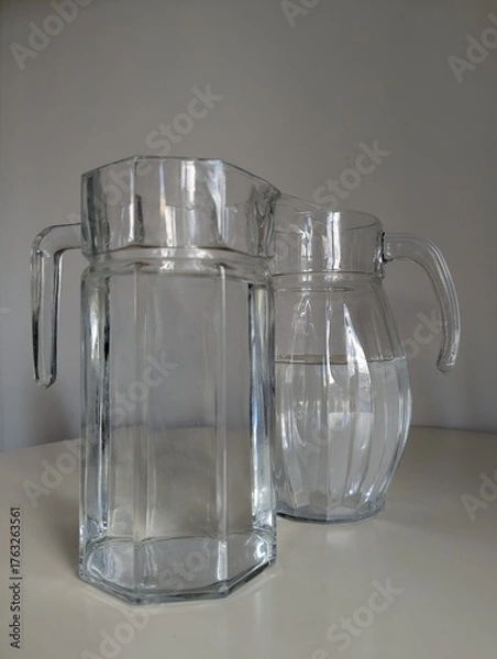 Fototapeta A minimalist duo of clear glass jugs with distinct shapes on a white table. Clean and modern aesthetic.