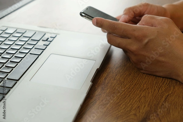 Fototapeta Person using smartphone near laptop on wooden desk, mobile transaction technology, digital communication and online connectivity concept