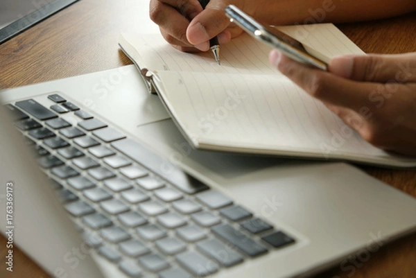 Fototapeta person holding smartphone while writing notes in notebook beside laptop, multitasking with focus and planning for business or study purposes