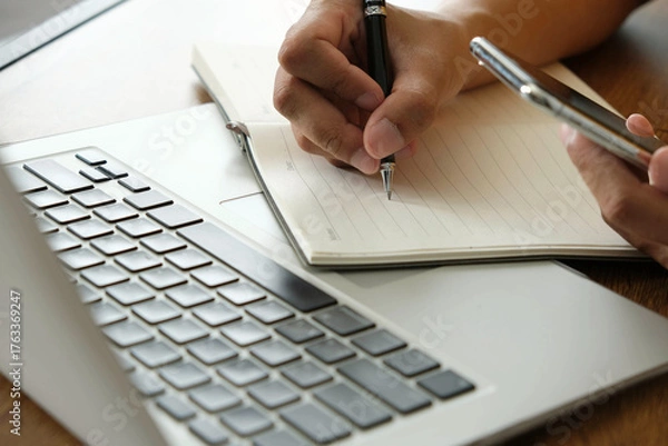 Fototapeta person writing notes in notebook while holding smartphone, with laptop keyboard in view, showing multitasking and business planning