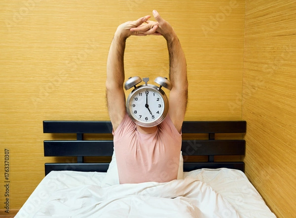 Fototapeta Happy man with alarm clock instead of head stretching on bed at home