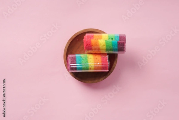 Fototapeta "A slice of rainbow roll cake sits on a wooden plate against a pink background."