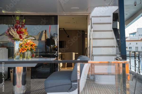 Obraz Elegant yacht interior with gray cushioned seating, modern table, glass railings, and fresh floral arrangements. Sunlight softly illuminates the cozy space, blending comfort with style.
