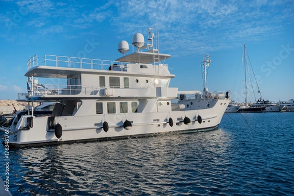 Fototapeta Large white yacht docked at marina. Multiple decks with railings and antennas on top. Other yachts visible in the background. Bright sunny day with reflections on the water  at Limassol Marina