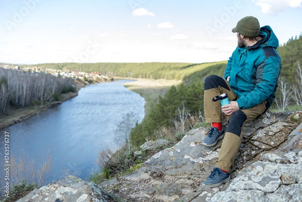 Fototapeta A bearded man is sitting resting on a rock with a thermocup in his hand, a knitted hat on his head