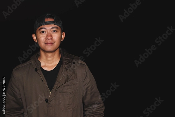 Fototapeta Young Asian Man Wearing Cap Portrait Against Dark Background with Copy Space. Studio-style portrait concept representing confidence, simplicity, and casual fashion for lifestyle or profile use.