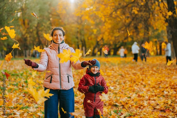 Fototapeta A woman and child joyfully play in a park amidst falling autumn leaves, surrounded by golden hues.