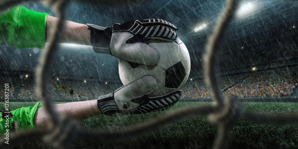Obraz Soccer goalkeeper catches a ball on big professional rainy football arena. View through the football goal. Goalkeeper wears unbranded sport uniform.
