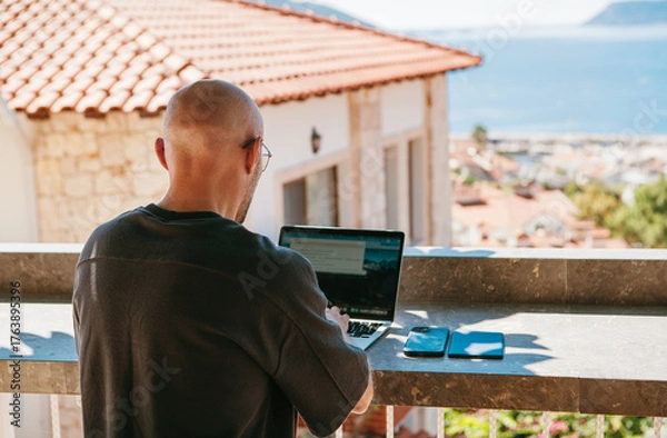 Fototapeta Freelancer bald fency man enjoying remote work on sunny terrace surrounded by greenery with sea view. Digital nomad using laptop embracing freedom flexible lifestyle in warm tropic island climate