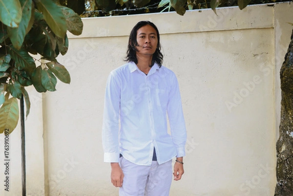 Fototapeta Serious Young Asian Man in White Shirt Standing Against Minimal Wall. He stands against a simple beige wall with leaves framing the top. Professional and minimalist style.